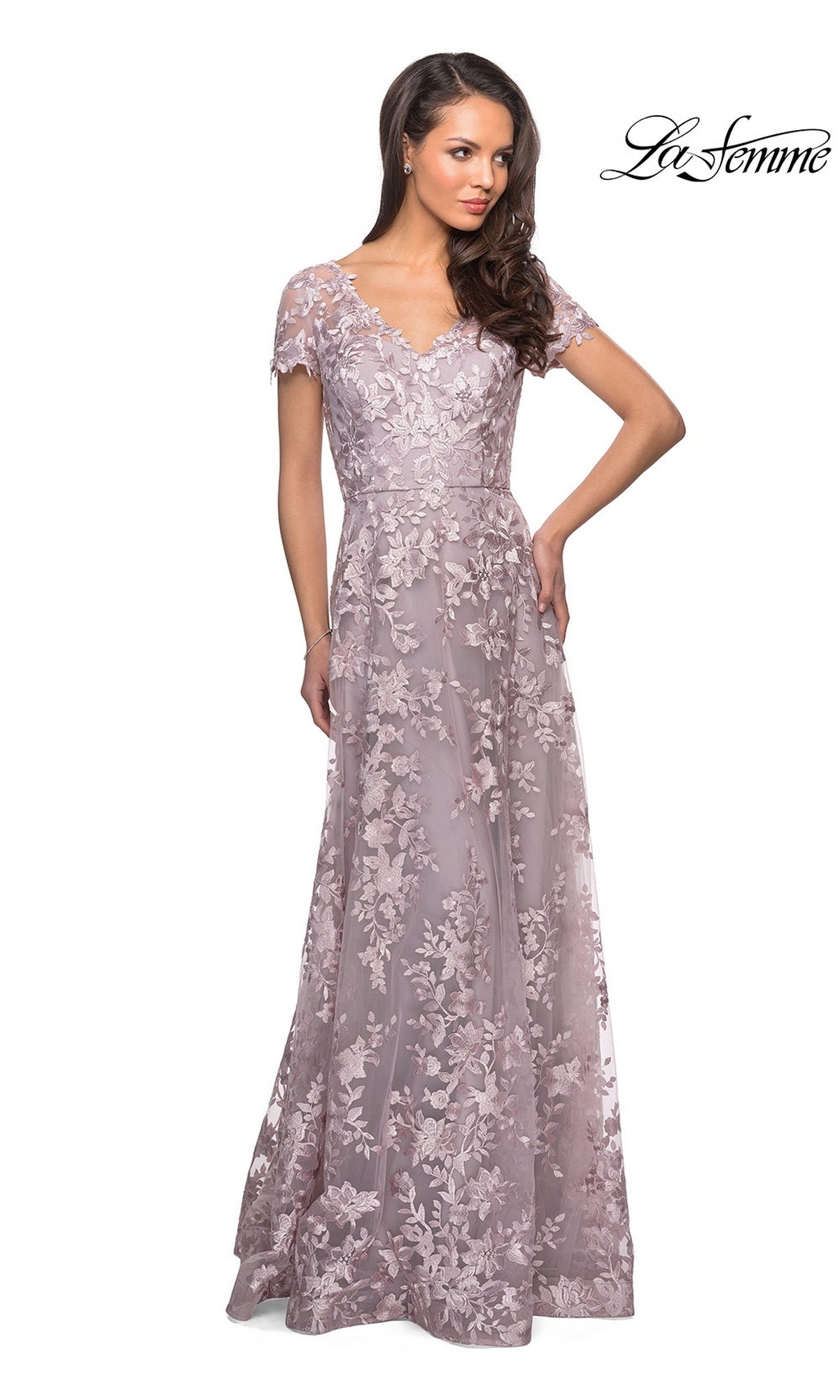 A woman poses with one hand on her hip, modeling the La Femme 27870—a floor-length, short-sleeved lavender lace mother of the bride dress adorned with floral patterns. The “La Femme” logo appears in the top right corner.